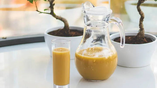 A glass and a jug of orange-colored juice sit on a glossy white table by a window. Two small potted plants with twisted trunks are in the background, adding greenery to the bright, serene setting.