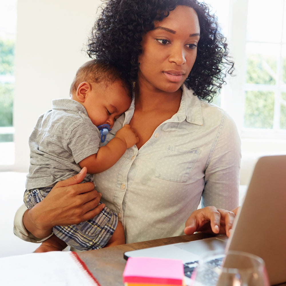 A woman works on a laptop at a desk while holding a baby who is sleeping on her chest. The desk has a notebook and colorful sticky notes, and sunlight streams in through a window behind them.