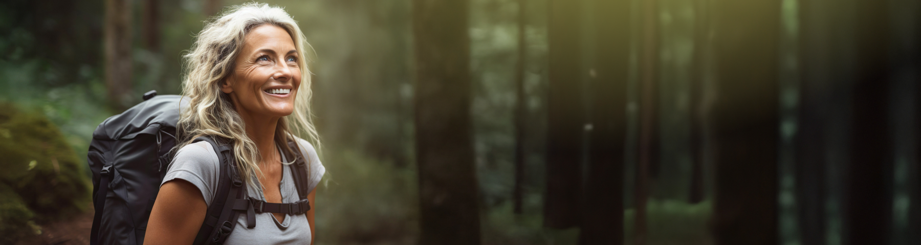 A woman with long blonde hair smiles while hiking in a forest, wearing a gray shirt and carrying a large backpack. Sunlight filters through the tall trees around her.