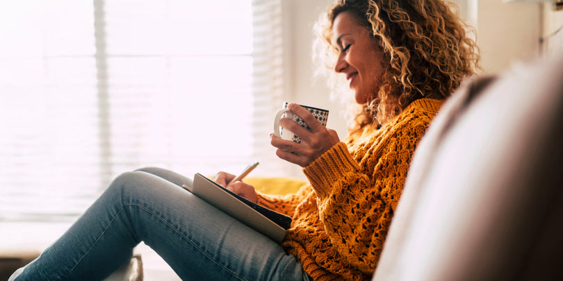 A woman with curly hair, wearing a yellow sweater and jeans, sits comfortably on a sofa, smiling as she writes in a notebook and holds a mug, with sunlight streaming through a window in the background.