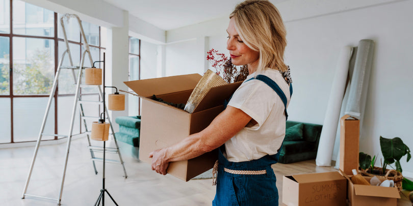 A woman holding a cardboard box stands in a bright, spacious room filled with moving boxes, a ladder, and rolled-up rugs, suggesting she is moving into or out of the space.