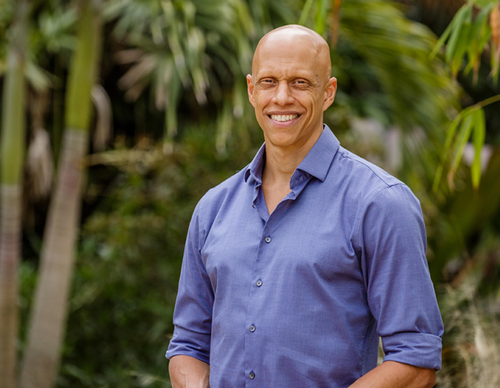 A smiling man stands outdoors, wearing a blue button-up shirt. The background features lush green foliage, including palm trees.