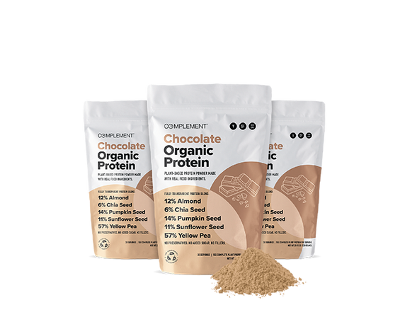 Three bags of Complement Chocolate Organic Protein powder are displayed. The packaging lists ingredients like almond, chia seed, and pumpkin seed. A small pile of protein powder is in front of the bags.