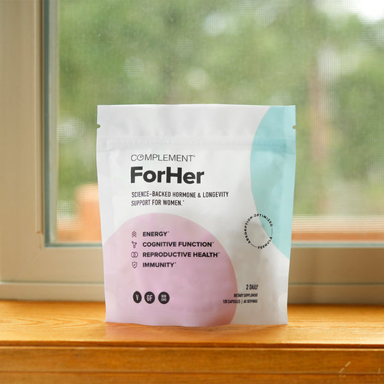 A pouch labeled Complement For Her sits on a wooden windowsill. The packaging highlights benefits including energy, cognitive function, reproductive health, and immunity. The design features pastel colors.