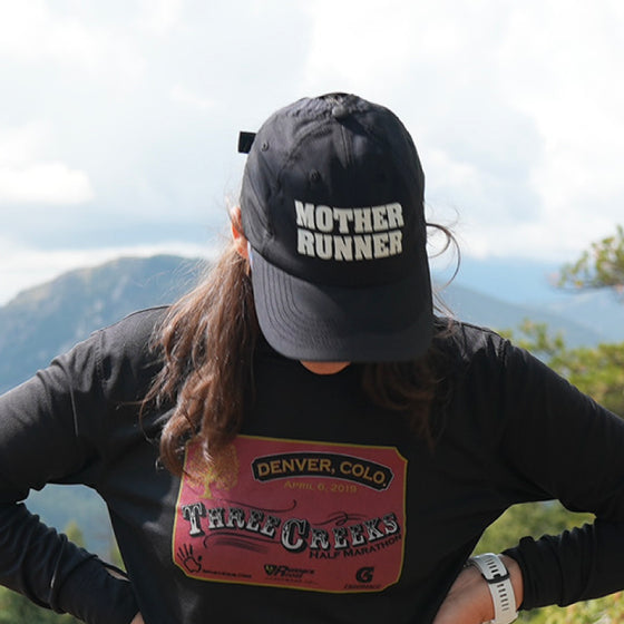 A person wearing a black cap with MOTHER RUNNER written on it and a long-sleeve shirt with a Denver, Colo Three Creeks Half Marathon logo. Theyre outdoors with a mountainous background.