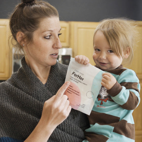A woman holds a young child in her arms. The child is holding a snack packet labeled ForHer. The woman appears to be looking at the packet with curiosity. The child is wearing a blue and brown outfit with a playful expression.