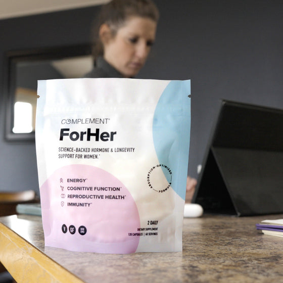 A woman sits at a table using a laptop. In the foreground, there is a pouch labeled Complement For Her, promoting energy, cognitive function, reproductive health, and immunity support for women.
