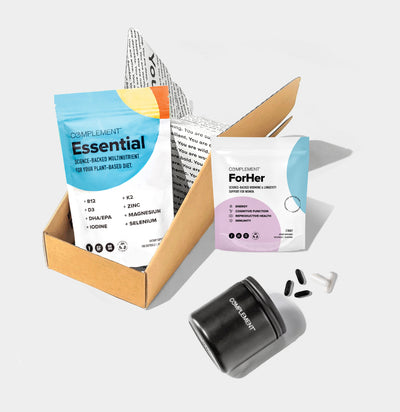 A set of health supplements displayed on a light background. Includes a cardboard box holding a blue and white package labeled Essential, a smaller package labeled For Her, and a black container with pills nearby.