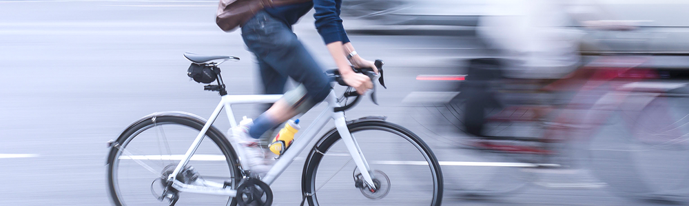 A person wearing jeans and a blue top rides a white bicycle quickly on a city street, with background cars and cyclists blurred to show motion.
