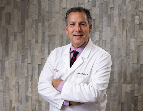 A man in a white lab coat with a maroon tie stands against a textured gray wall. He is smiling with arms crossed, exuding confidence and professionalism.