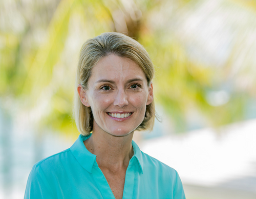A woman with short blonde hair smiles while wearing a teal blouse. She stands outdoors with a blurred background of green foliage and bright sunlight.