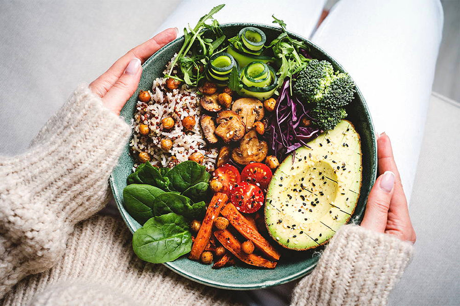 A person holding a bowl of healthy food, including avocado, cherry tomatoes, chickpeas, spinach, arugula, broccoli, quinoa, mushrooms, and carrots. The person is wearing a cozy beige sweater.