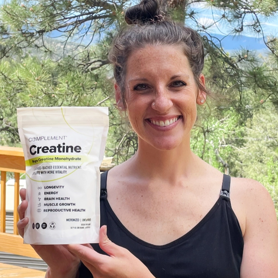 A person smiling and holding a bag of Complement Creatine Monohydrate powder. They are standing outdoors on a deck, with trees and mountains in the background.