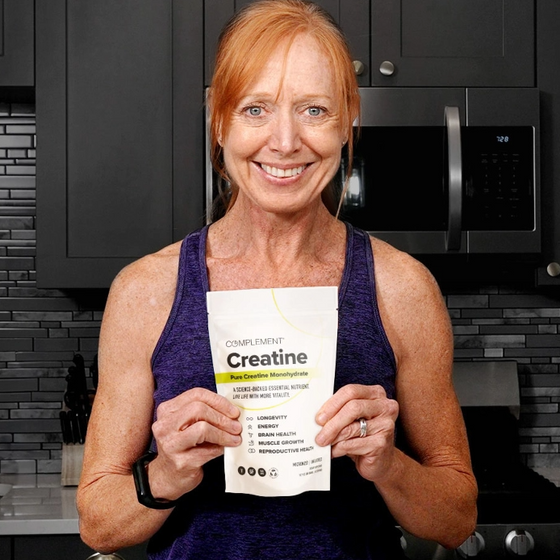A woman in a kitchen holds a bag of Creatine supplement. She is wearing a purple tank top and has red hair. Kitchen appliances and a tile backsplash are visible in the background.