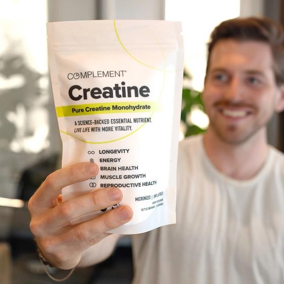 A person holds a white package labeled Complement Creatine with text highlighting benefits such as longevity, energy, brain health, muscle growth, and reproductive health. The background is slightly blurred, focusing on the product.