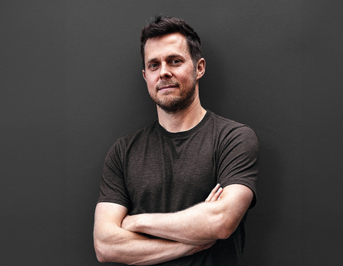 A man with short dark hair and facial hair stands with arms crossed against a dark gray background. He is wearing a simple dark t-shirt and has a neutral expression.