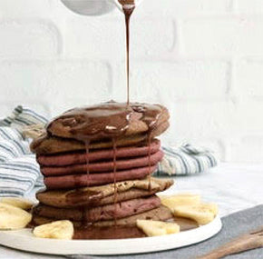 A stack of chocolate pancakes on a white plate, topped with chocolate syrup being poured over them. Slices of banana are arranged around the base. A striped cloth is in the background.