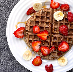 A plate with a circular waffle topped with sliced strawberries and bananas, drizzled with syrup, placed on a gray surface.