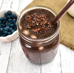 A mason jar filled with a thick, dark purple smoothie topped with cocoa nibs and a straw. The jar is placed on a wooden surface with a bowl of blueberries in the background.