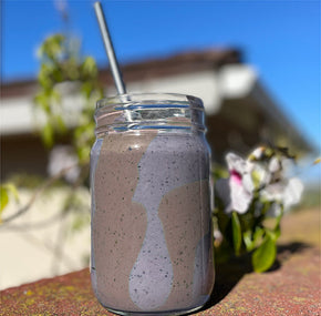 A mason jar filled with a creamy purple smoothie, topped with a metal straw. The jar is placed on a ledge with a blurred background of greenery and a building under a clear blue sky.