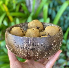 A hand holds a coconut bowl containing several chocolate chip cookie dough balls. The bowl has the phrase Good Food, Happy Mood printed on it. Green foliage is blurred in the background.