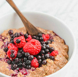 A bowl of oatmeal topped with fresh raspberries and blueberries. A wooden spoon is placed in the bowl.