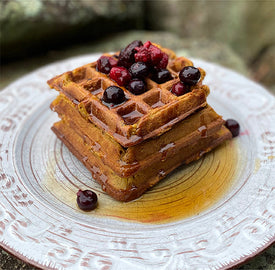 A stack of golden-brown waffles topped with syrup and mixed berries, served on a decorative white plate. The background is softly blurred, emphasizing the breakfast dish.
