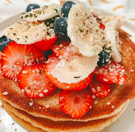 A stack of pancakes topped with sliced strawberries, blueberries, banana slices, and sprinkled with seeds, served on a white plate.