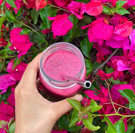 A hand holding a glass jar with a bright pink smoothie and a straw, surrounded by vibrant pink bougainvillea flowers.