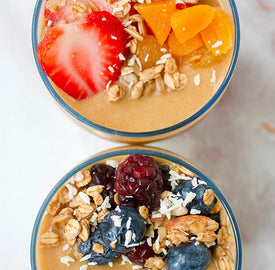 Two smoothie bowls topped with fruit and granola. The top bowl features sliced strawberries, peaches, and granola. The bottom bowl includes blueberries, blackberries, granola, and shredded coconut. Both bowls are on a light surface.