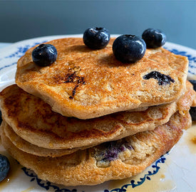 A stack of three blueberry pancakes on a white and blue plate. The pancakes are golden brown with visible blueberries inside, and there are several fresh blueberries on top.