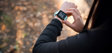 A person outdoors wearing a black long-sleeve top checks fitness stats on a smartwatch with a light blue strap, surrounded by a blurred, natural background.