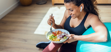 A woman in workout clothes sits on the floor next to a blue exercise ball, smiling and eating a colorful salad from a bowl. She is wearing wireless earbuds and appears relaxed and happy.