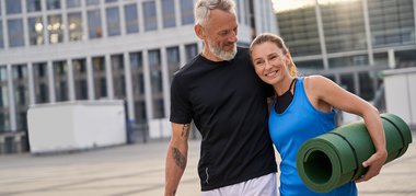 Smiling middle-aged couple in sportswear stands outdoors; the woman holds a rolled-up yoga mat and leans on the man as they look at each other, with modern buildings in the background.
