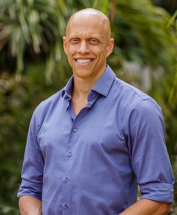 A smiling bald man wearing a light blue button-up shirt stands outdoors in front of leafy green plants.
