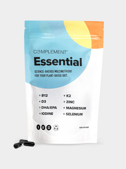 A white and colorful resealable pouch labeled Complement® Essential by Complement features a list of nutrients like B12, D3, K2, omega-3 DHA/EPA, magnesium, and selenium, with two black capsules shown beside the package.