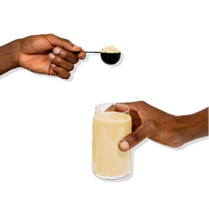 A hand holds a black scoop of protein powder above another hand holding a glass of beige protein shake. The background is white, and the focus is on the powder and the drink.