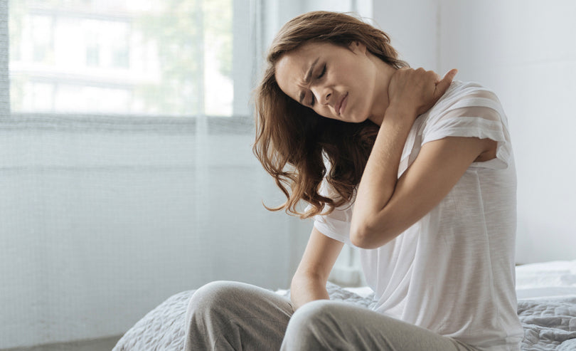 A woman sitting on a bed holds her neck with one hand and winces in pain. She is wearing a white t-shirt and gray pants in a softly lit room with light-colored curtains.