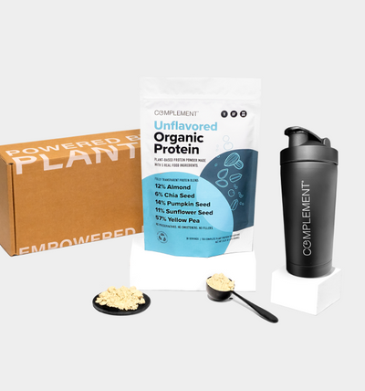 A protein powder package labeled Complement Unflavored Organic Protein stands between a cardboard box and a black shaker bottle. A scoop and mound of powder are in front, with text on the package listing almond, chia, pumpkin, sunflower, and yellow pea.