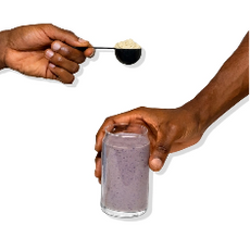 A hand holds a scoop of protein powder over a glass of purple smoothie, which another hand is grasping. The background is plain white.