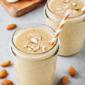 Two glasses of almond banana smoothie are topped with sliced almonds and have striped straws. Surrounded by whole almonds, the drinks sit on a light gray surface with a wooden board in the background.