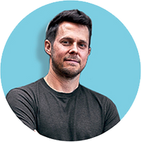 A man with short dark hair and a beard stands against a light blue background, wearing a dark gray t-shirt and smiling slightly.