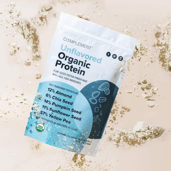 A bag of Complement Unflavored Organic Protein powder is surrounded by floating protein powder. The packaging highlights its plant-based blend: almond, chia seed, pumpkin seed, sunflower seed, and yellow pea.