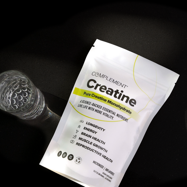 A white packet labeled Creatine Pure Creatine Monohydrate lies on a dark surface next to a glass of water. The packet highlights benefits like energy, brain health, muscle growth, and reproductive health.