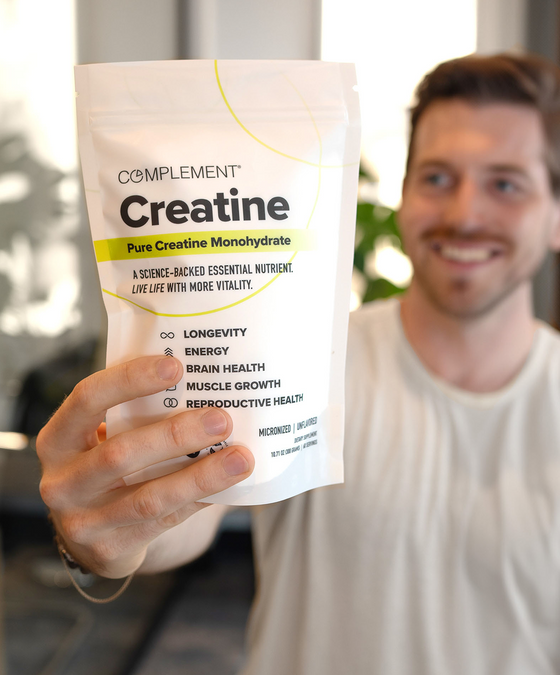 A smiling man holds up a bag of Complement Creatine supplement, showing the front label that highlights benefits like longevity, energy, brain health, muscle growth, and reproductive health.