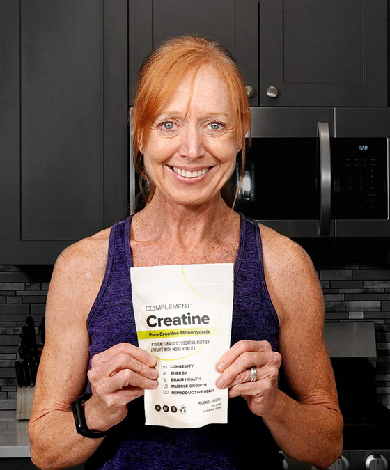 A smiling woman with red hair in a ponytail, wearing a purple tank top, stands in a kitchen holding a packet of Complement Creatine supplement in front of her chest.