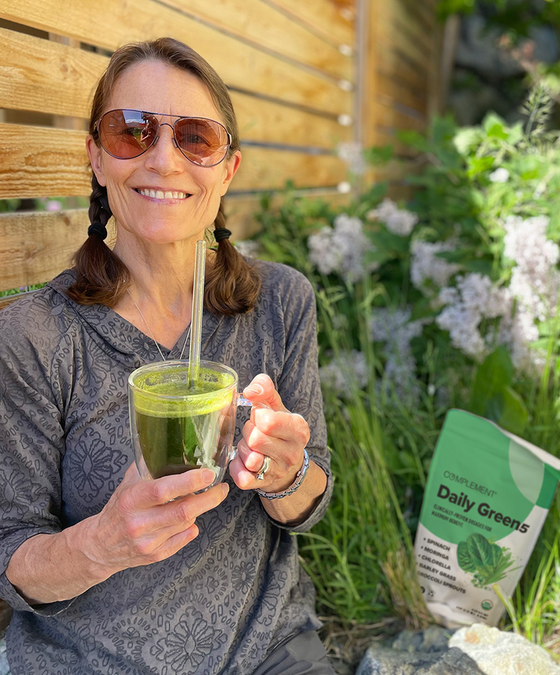 A smiling woman with sunglasses and braided hair sits outdoors holding a glass of green smoothie. A bag labeled “Daily Greens” is on the ground beside her. Lush greenery and a wooden fence are in the background.