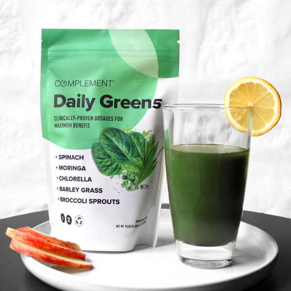 A glass of green smoothie with a lemon slice sits next to a Complement Daily Green 5 supplement bag, featuring ingredients like spinach, moringa, chlorella, barley grass, and broccoli sprouts. Apple slices are also nearby.