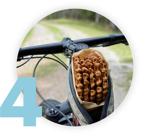 A close-up of a bikes handlebar and frame bag containing pretzels. A large blue number 4 is in the lower left corner. The bike is on a path surrounded by greenery.