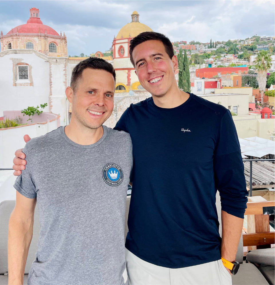 Two men smiling with their arms around each other on a rooftop. They are in casual clothing, and the background features historic buildings with colorful domes under a cloudy sky.